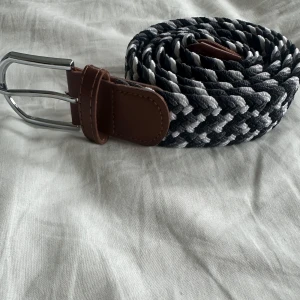 Svart, vit och grå färjat bälte  - Snyggt flätat bälte i svart, vitt och grå med bruna skinn-detaljer vid spännet och änden. Bältet har ett coolt randigt mönster och ett silvrigt metallspänne som ger en modern vibe.