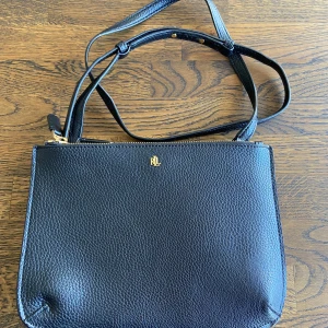 Svart väska från Ralph Lauren - Stilren svart axelväska från Ralph Lauren med justerbar rem och guldfärgad RL-logga framtill. Tvådelat innerfack Förslutning: Dragkedja Utvändigt material: Skinn Foder: Polyester. En liten skav på axelremmen, se bild.