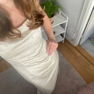 Champagnefärgad långklänning med snörning - Elegant långklänning i champagnefärg med tunna axelband och vacker broderad dekor framtill. Klänningen har snörning och knappar i ryggen för en snygg passform. Materialet har en lätt glans och känns lyxigt, perfekt för speciella tillfällen.