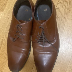 Bruna snörskor från H&M i skinnimitation - Stilrena bruna snörskor från H&M i skinnimitation med spetsig tå och platt sula. Perfekta för en dressad look med klassisk design och diskreta sömmar. Skorna har mörkbruna snören och svart innersula.