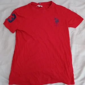 Röd USPA t-shirt med tryck - Röd t-shirt från US Polo Assn med blått polospelare-tryck på bröstet och en stor blå siffra 3 på ena ärmen. Klassisk rund hals och korta ärmar. Skön och enkel modell i bomull, perfekt för en sportig och avslappnad look.