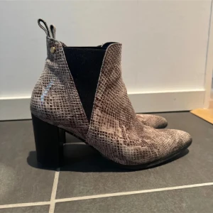 Bruna stövletter med ormmönster - Snygga bruna stövletter med chunky klack och ormmönstrad yta. Skorna har rund tå och elastiska svarta paneler på sidorna för enkel påtagning. Perfekt för dig som vill sticka ut med coola boots i höst och vinter.