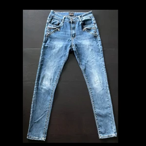 Blå skinny jeans Chica London - Snygga blå skinny jeans från Chica London med coola dragkedjor och dekorativa detaljer vid fickorna. Jeansen har normal midja och klassisk femficksdesign. Perfekta för dig som gillar en smal passform och vill sticka ut lite extra.