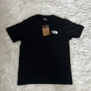 Svart t-shirt från The North Face - Svart t-shirt från The North Face med klassisk logga tryckt i vitt på bröstet. Enkel och stilren design med rund hals och korta ärmar. Perfekt för dig som gillar streetwear och vill ha en clean look. Materialet känns mjukt och bekvämt mot huden.