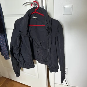 Svart vindjacka från H&M Sport - Svart vindjacka från H&M Sport i storlek M. Jackan har dragkedja framtill, hög krage och resår vid ärmslut och midja. 