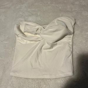 Vit bandeau tubtopp med twist - Supersnygg vit bandeau tubtopp med twistad detalj framtill. Toppen är ribbad och axelbandslös, perfekt för varma dagar eller när du vill ha en clean look. Mjuk och stretchig, sitter skönt och formar sig efter kroppen.