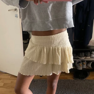 Krämvit volangkjol med struktur - Supersöt kort kjol i krämvitt med två lager volanger och smockad midja. Kjolen har en skön, strukturerad känsla och är perfekt för dig som gillar en feminin och trendig stil. Passar lika bra till en enkel hoodie som till en snygg topp.