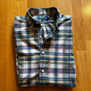 Ralph Lauren skjorta - Snygg rutig skjorta från Polo Ralph Lauren i slim fit. Skjortan har blå, grön, gul och röd färg med klassiskt broderat rött logomärke på bröstet. Tillverkad i mjuk bomull och har knappar framtill samt button down-krage. Hör av dig vid funderingar. Mvh Carl