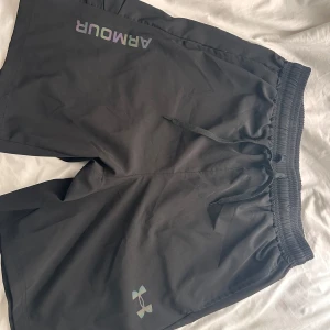Svarta träningsshorts från Under Armour - Svarta träningsshorts från Under Armour med elastisk midja och snörning. Shortsen har en diskret regnbågsfärgad logga och text på ena benet. Perfekta för gymmet eller löprundan, med en loose fit och sportig känsla.
