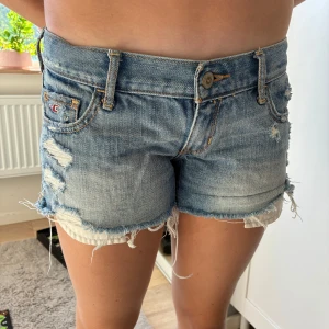 Blå slitna jeansshorts från Hollister - Snygga blå jeansshorts från Hollister med slitna detaljer och fransig kant. Klassisk femficksmodell med låg midja och coola slitningar både fram och bak. Ikonisk Hollister-logga på fickan och bak. Jag har fler bilder om någon undrar!