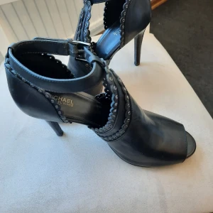 Svarta pumps från Michael Kors - Snygga svarta pumps i skinn från Michael Kors med öppen tå, hög klack och coola detaljer med nitar och vågig kant. Rem runt ankeln med spänne ger extra edge. Perfekta för dig som vill sticka ut med en stilren men ändå unik look.