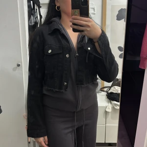 Svart croppad jeansjacka - Säljer en svart croppad jeansjacka med rå kant nertill och silvriga knappar. Jackan har klassisk krage, två bröstfickor och långa ärmar. Perfekt för dig som vill ha en edgy och trendig look till vardags.