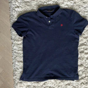 Mörkblå pikétröja från Morris - Säljer en klassisk mörkblå pikétröja från Morris med liten röd broderad logga på bröstet. Tröjan har korta ärmar, krage och knappar framtill. Perfekt för en stilren och avslappnad look. Nypris 999kr 