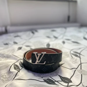 Lv bälte  - Säljer ett svart bälte (HELT NYTT) från Louis Vuitton med klassiskt monogrammönster och ett stort silverfärgat LV-spänne. Snyggt Lv bälte sitter riktigt bra. Storlek 105cm. Aldrig använt. Bältet är gjort i skinn och har en brun insida med präglad märkning. Perfekt accessoar för att lyfta din outfit. Tveka inte hör av dig vid frågor