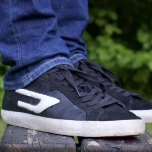 Svarta sneakers från Diesel med vit detalj - Säljer ett par svarta sneakers från Diesel med vit sula, vit häl och vit logodetalj på sidan. Skorna har snörning och en röd undersida på sulan. Perfekta för en avslappnad stil och passar till jeans eller shorts.