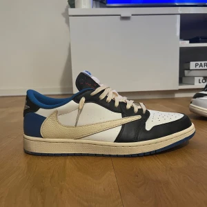 Nike Air Jordan 1 Low OG Travis Scott x Fragment - Nike Air Jordan 1 Low OG Travis Scott x Fragment med vit, svart och blå färg. Skorna har omvänd swoosh i beige, svarta och blå detaljer samt klassisk snörning. Perfekta för dig som gillar unika sneakers med streetwear-känsla.