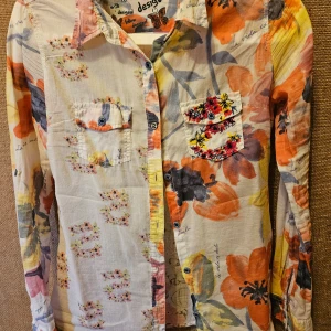 Färgglad blommig skjorta från Desigual i str. S - Säljer en tunn och färgglad skjorta från Desigual med stora blommor i orange, gul, svart och grått. Skjortan har lång ärm, knappar framtill och två bröstfickor, varav en är broderad med blommor och bin. Perfekt för dig som gillar unika plagg!