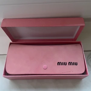 Svarta solglasögon från Miu Miu - Stilrena svarta solglasögon från Miu Miu med markerad, rektangulär båge och guldiga detaljer på sidorna. Kommer med ett rosa fodral med tryckknapp och Miu Miu-logga. Perfekt accessoar för att lyfta din look.