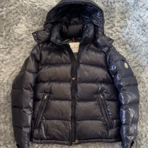 Svart pufferjacka från Moncler - Säljer en svart pufferjacka från Moncler med huva och dragkedja framtill. Jackan har två snedställda fickor med dragkedja och Moncler-logga på ärmen. Klassisk glansig finish och quiltad design.