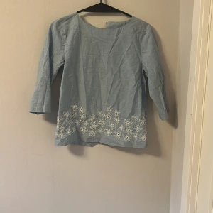 Ljusblå blus med broderade blommor från GANT - Säljer en ljusblå blus från GANT med trekvartsärm och vita broderade blommor längst kanten. är tillverkad i ett tunt, svalt material. Perfekt för vår och sommar!