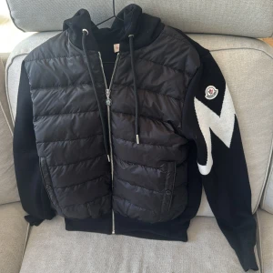 Moncler Jacka Cardigan  - Säljer en svart jacka från Moncler med huva och dragkedja. Jackan har quiltad framsida, två stora fickor och en vit Moncler-logga samt mönster på ärmen. Perfekt för dig som vill ha en snygg och sportig look. Funkar båda för våren/sommaren Storlek Medium, men passar även Small då jag är S/M och passar mig. Säljs för 2500kr