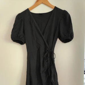 Svart omlottklänning med puffärm från Gina Tricot - Säljer en svart omlottklänning från Gina Tricot med puffiga kortärmar och volang nertill. Klänningen har knyt i midjan och en feminin siluett, perfekt för dig som gillar stilrena plagg med fina detaljer.