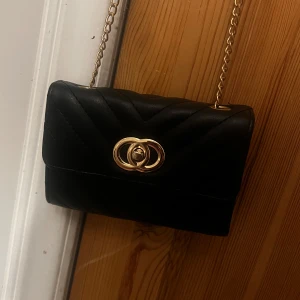 Svart quiltad axelväska med guldkedja - Säljer en svart quiltad axelväska med guldfärgad kedja och snyggt spänne framtill. Väskan har flera fack inuti, inklusive ett med dragkedja. Perfekt storlek för det viktigaste och ger en stilren look.