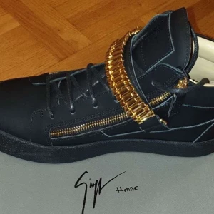 Svarta sneakers med gulddetaljer från Giuseppe Zanotti - Unika svarta sneakers med guldfärgade dragkedjor och en bred guldfärgad rem över vristen som liknar en klocklänk. Skorna har rund tå och platt sula, samt snörning framtill. Perfekta för dig som vill sticka ut med exklusiva detaljer.
