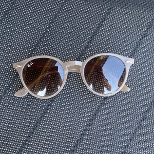 Beige runda solglasögon från Ray-Ban - Snygga runda solglasögon från Ray-Ban i beige plast med bruna glas. Modellen är RB 2180 och har en klassisk, trendig look med Ray-Ban-logga på bågen. Perfekta för soliga dagar och ger en cool touch till din outfit.