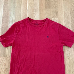 Röd t-shirt från Polo Ralph Lauren - Röd t-shirt från Polo Ralph Lauren med klassisk passform och liten blå broderad logga på bröstet. Perfekt basic-plagg med rund hals och korta ärmar. Skön och mjuk bomullskvalitet.