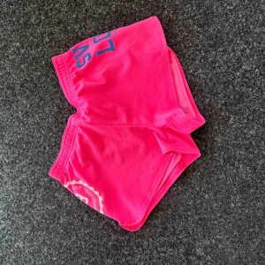 Rosa sportiga shorts från SVEA - Säljer ett par knallrosa sportshorts från SVEA med tryck i vitt och blått. Shortsen har resår i midjan och är tillverkade i ett syntetmaterial som andas. Perfekta för träning eller sommarens varma dagar.