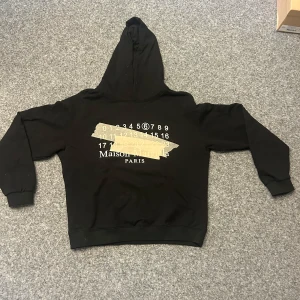 Svart hoodie från Maison Margiela - Säljer en svart hoodie från Maison Margiela Paris med ikoniskt siffror-tryck och beige tejpdetaljer på bröstet. Tröjan har huva och ribbade muddar vid ärmslut och nederkant. Perfekt för dig som gillar streetwear och unika designer.
