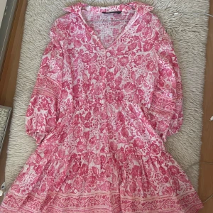 Rosa och vit blommig klänning med volang - Superfin klänning i rosa och vitt med blommigt mönster och volangdetaljer. Klänningen har v-ringning och trekvartsärmar, perfekt för sommarens alla tillfällen. Lätt och luftig modell som ger en romantisk känsla.