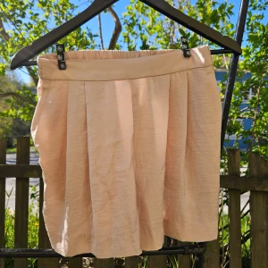Beige/rosa shorts - Säljer söta rosa shortsl med bred linning och fina veck framtill. Shotsen har en lätt och luftig känsla, perfekt för varmare dagar. Enkel att matcha med olika toppar för en fräsch look.