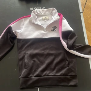 Svart, vit och rosa tröja från Illusive London - Säljer en sportig tröja från Illusive London i svart, vitt och rosa. Tröjan har halv dragkedja, hög krage och fickor framtill. Snygg detalj med rosa rand längs ärmen och broderad logga på bröstet.