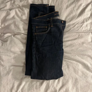 Mörkblå jeans från Nudie Jeans - Säljer ett par mörkblå jeans från Nudie Jeans med klassiska orange sömmar och snygga bakfickor med vågigt broderi. Jeansen har normal passform och är tillverkade i bomull. Perfekta för dig som gillar stilrena och tidlösa jeans. Jeansen är andvända 3 GÅNGER totalt detta eftersom jag fick dem i present men gillade inte modellen på dem. 