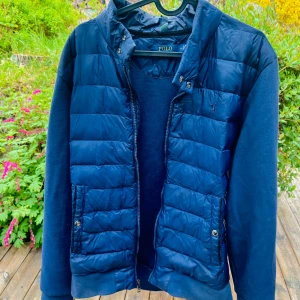 Blå pufferjacka från Polo Ralph Lauren - Snygg blå pufferjacka från Polo Ralph Lauren med dragkedja och quiltade partier framtill. Jackan har långa ärmar i mjukt material och två fickor med knappar. Perfekt för kyliga dagar.