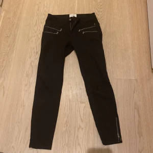 Svarta byxor med dragkedjedetaljer från Freequent - Svarta byxor från Freequent med smal passform och flera dragkedjor framtill samt vid bensluten. Byxorna har en klassisk midja och raka ben. Perfekta för en stilren och modern look.