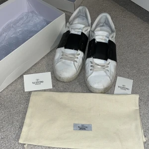Valentino Garavani Open sneakers med svart detalj - Säljer ett par vita Valentino Garavani Open sneakers med bred svart läderdetalj över vristen. Klassisk rund tå, platt sula och snörning. Skorna kommer med originalkartong, dustbag och äkthetskort. Kan gå ner i pris vid snabb affär. Har inte orkat tvätta dem så dem kan bli mycket fräschare, säljer pga att jag vill ha lite nya golfklubbor.