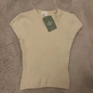Beige ribbad stickad topp från H&M - Säljer en beige ribbad stickad topp från H&M. Toppen har korta ärmar och är figurnära med rund halsringning. Tillverkad av återvunnet material, perfekt för en stilren vardagslook.