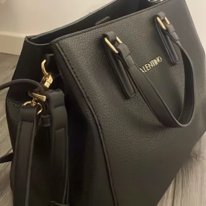 Svart handväska från Valentino - Snygg svart handväska från Valentino med guldfärgade detaljer och dubbla handtag. Väskan har axelrem och dragkedja upptill samt stilren design i strukturerat skinnliknande material. Perfekt för att lyfta din outfit.