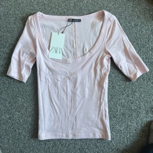 Ljusrosa topp från Zara - Säljer en ljusrosa topp från Zara med djup rundad urringning och korta ärmar. Toppen har en tight passform och är tillverkad i ett mjukt, stretchigt material. Perfekt att styla till jeans eller kjol!