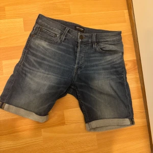 Blå jeansshorts från Jack & Jones - Snygga blå jeansshorts från Jack & Jones med klassisk femficksdesign och uppvikta benslut. Shortsen har normal passform och stängs med dragkedja och knapp. Perfekta för varma dagar och enkel att matcha med t-shirt eller skjorta.