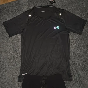 Svart t-shirt från Under Armour med regnbågsdetaljer - Säljer en svart t-shirt från Under Armour med färgglada loggor och textdetaljer i regnbågsfärger på bröstet, ärmen och sidan. T-shirten har rund hals och korta ärmar, perfekt för träning eller sport.