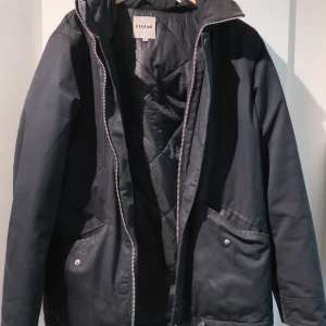 Svart jacka från Elvine med huva - Svart jacka från Elvine med huva och dragkedja framtill. Jackan har två stora fickor med lock och knapp, samt en bröstficka med dragkedja. Perfekt för kyligare dagar och har en stilren, enkel design.