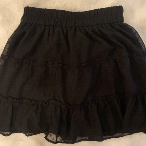 Svart volangkjol från New Yorker  - Supersöt svart kortkjol från New Yorker med volangdetaljer och resår i midjan. Kjolen har ett diskret prickigt mönster i tyget och är perfekt för dig som gillar en feminin och trendig stil.