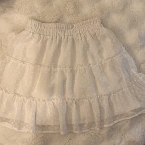 Vit volangkjol med prickigt mönster från från New Yorker  - Supersöt vit kortkjol från New Yorker med volanger och diskret prickigt mönster. Kjolen har resår i midjan och är gjord i ett lätt, luftigt material som passar perfekt till varmare dagar. 