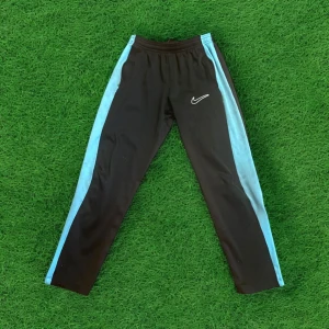 Svarta träningsbyxor från Nike med blåa sidor - Snygga svarta träningsbyxor från Nike med ljusblåa paneler längs sidorna och vit broderad logga på låret. Byxorna har elastisk midja och är perfekta för sport eller chill. Klassisk sportig look!