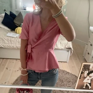 Rosa omlottblus  - Säljer en söt rosa omlottblus med knyt i midjan och korta ärmar i storlek M (passar perfekt för S också). Blusen har en v-ringad design och markerad midja som ger en fin siluett. Perfekt att styla med jeans eller kjol. (Har även lapp kvar)