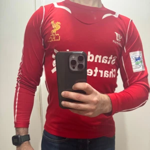 Liverpool långärmad fotbollströja - Röd långärmad Liverpool-tröja med vita detaljer och klubbmärke på bröstet. Tryckt sponsorlogga på framsidan och broderad gul Liverbird. Perfekt för fans som vill visa sitt stöd.